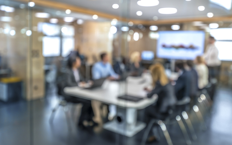 Blurred view of a business team meeting around a table in a glass-walled conference room.